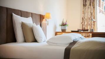 A neatly made bed with white linens and a brown headboard, a bedside lamp, and a small table with a potted plant in the background.