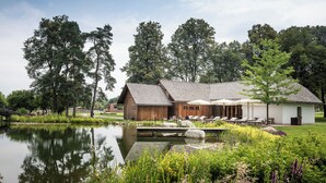 Un bâtiment rustique en bois, avec un étang devant, entouré d’une végétation luxuriante et d’arbres.