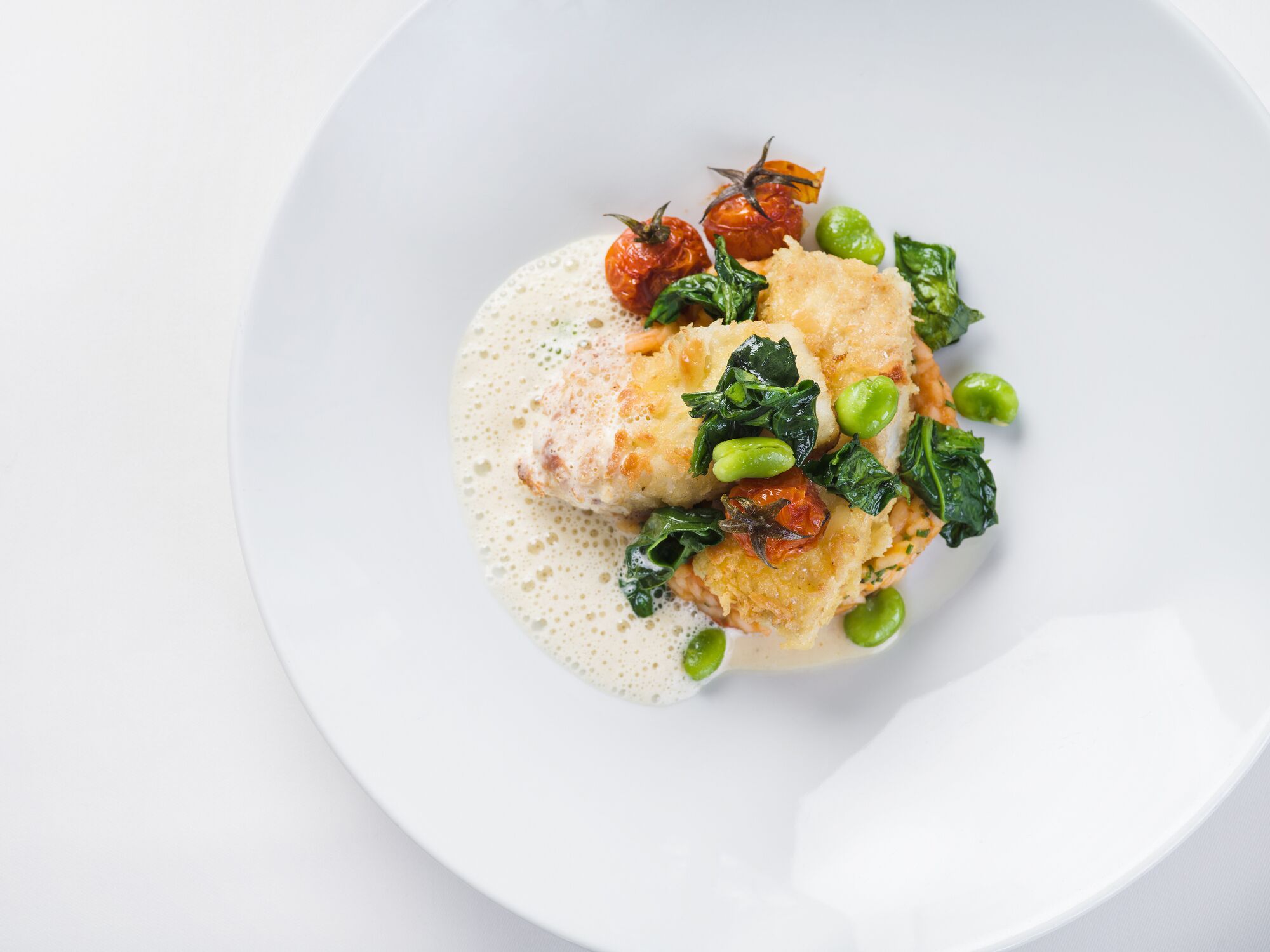 Ein weißer Teller mit einer Portion Essen, garniert mit Erbsen, einer Kirschtomate und Blattgemüse, dazu eine cremige Soße.