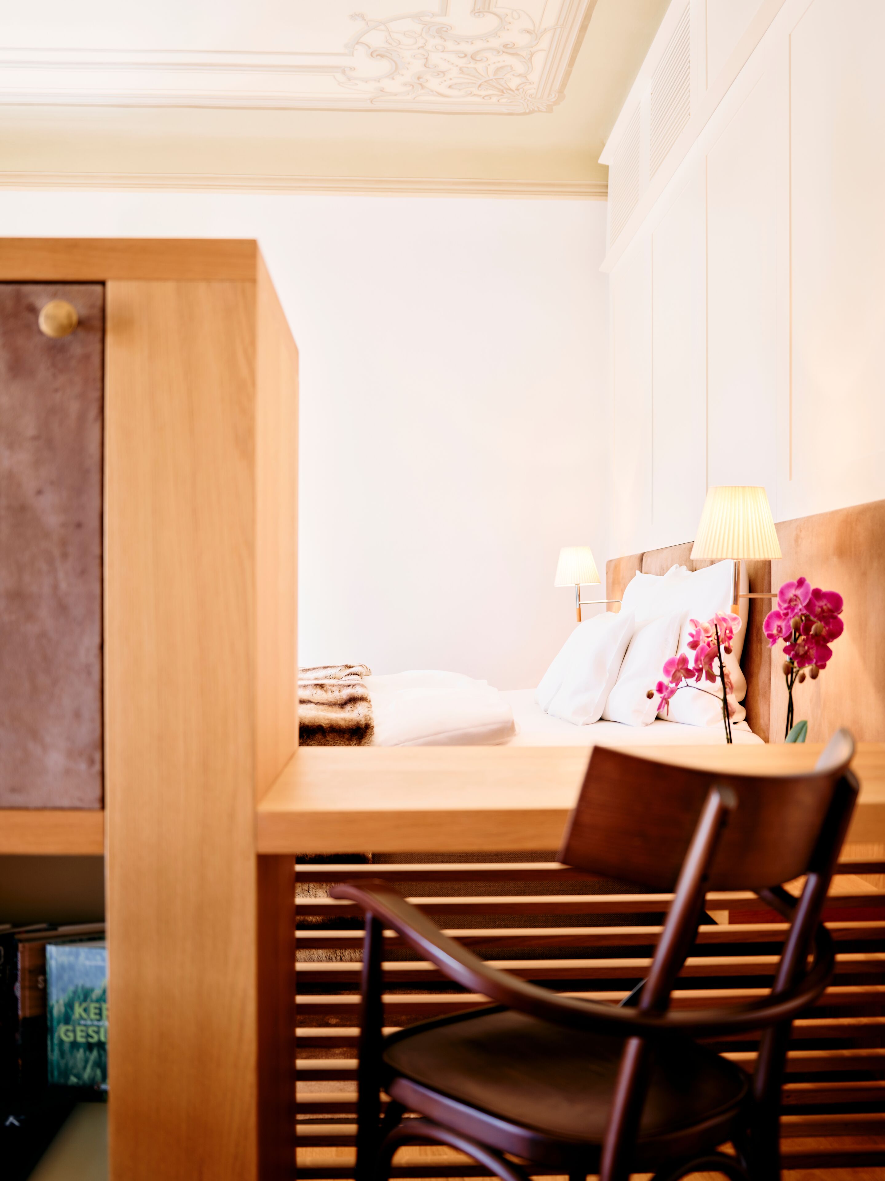 A modern hotel room with a wooden desk, a chair, a bed with white linens, a lamp, and a vase with flowers.