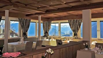 Ein Restaurant mit mehreren Tischen, großen Fenstern mit Vorhängen und Blick auf ein Gebirge.