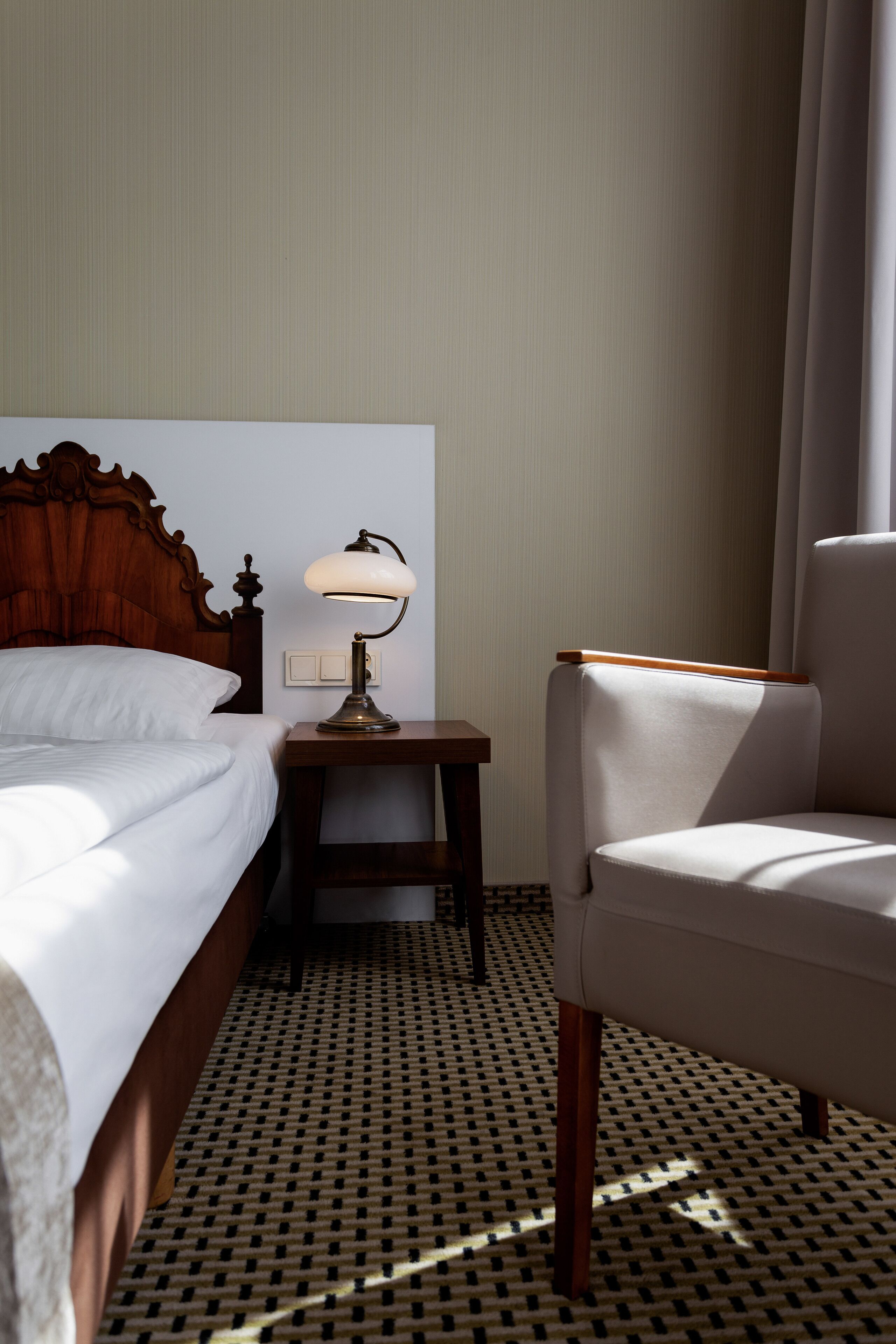 A hotel room with a wooden headboard bed, a bedside lamp, a small table, and a chair.