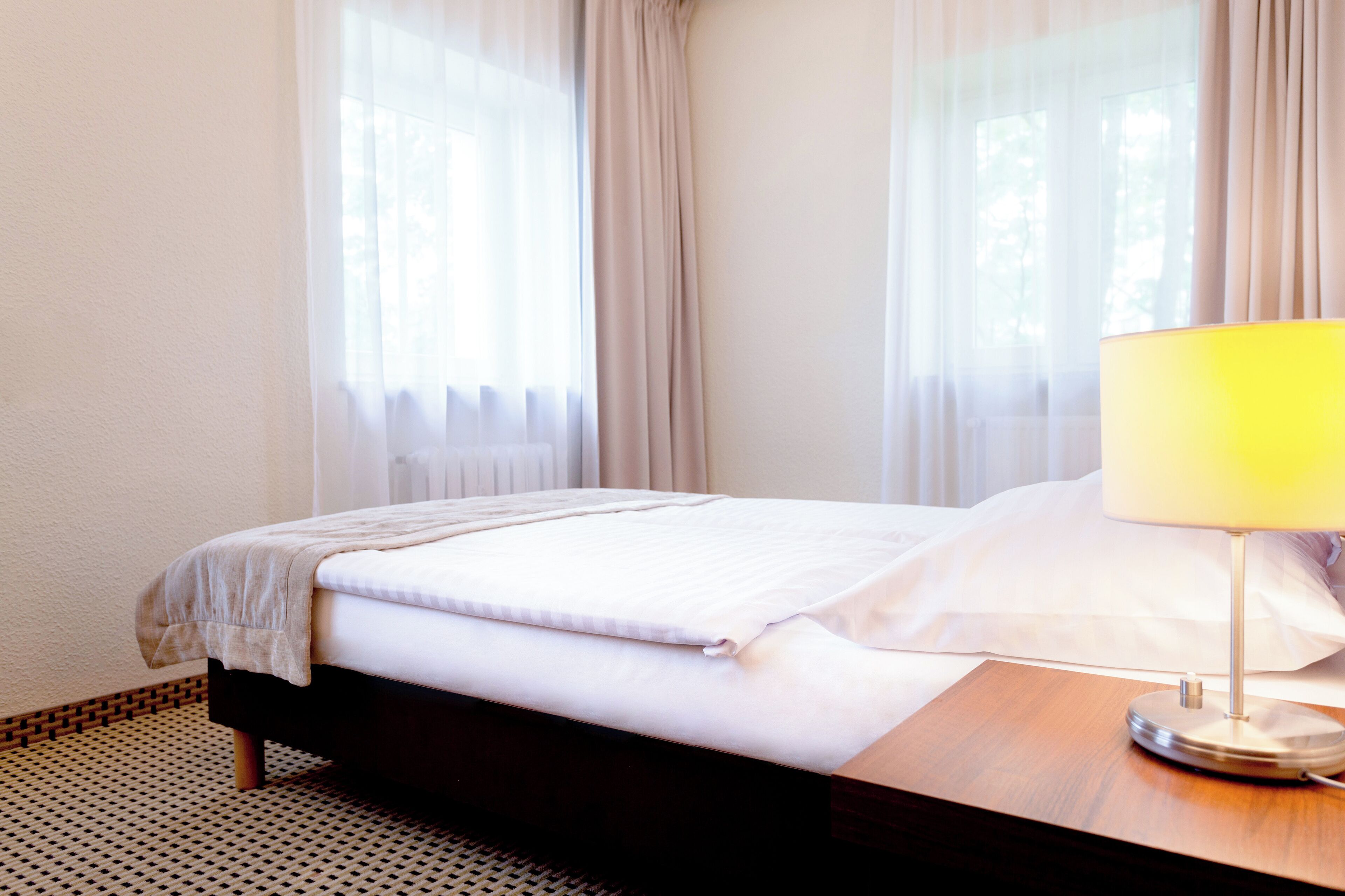 Una cama bien tendida con edredón blanco y manta de color beige, una mesita de noche de madera con lámpara, y cortinas traslúcidas en una ventana.