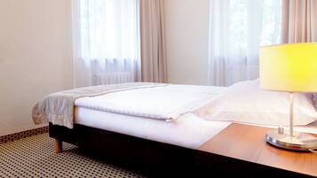 Una cama bien tendida con edredón blanco y manta de color beige, una mesita de noche de madera con lámpara, y cortinas traslúcidas en una ventana.