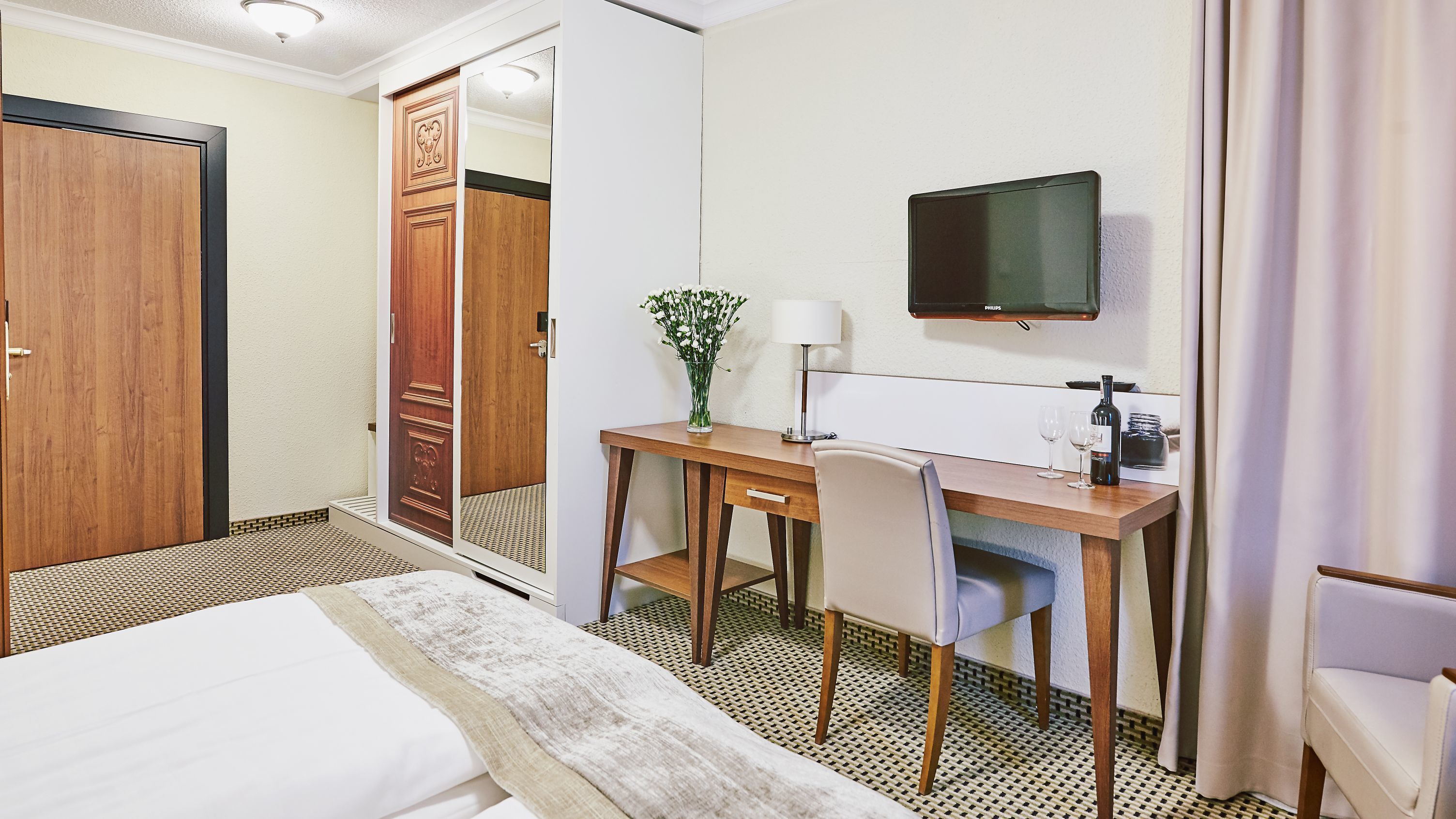 A hotel room with a bed, a desk with a chair, a TV, and a small table with a vase of flowers.