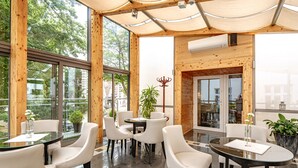Un espacio interior moderno con amplios ventanales, paredes de madera y una combinación de mesas redondas y cuadradas con sillas blancas.