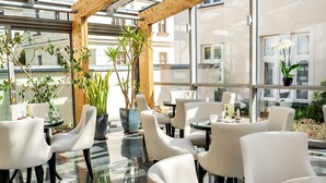 Un comedor moderno con mesas de vidrio y sillas blancas, rodeado de ventanas amplias y plantas en macetas.