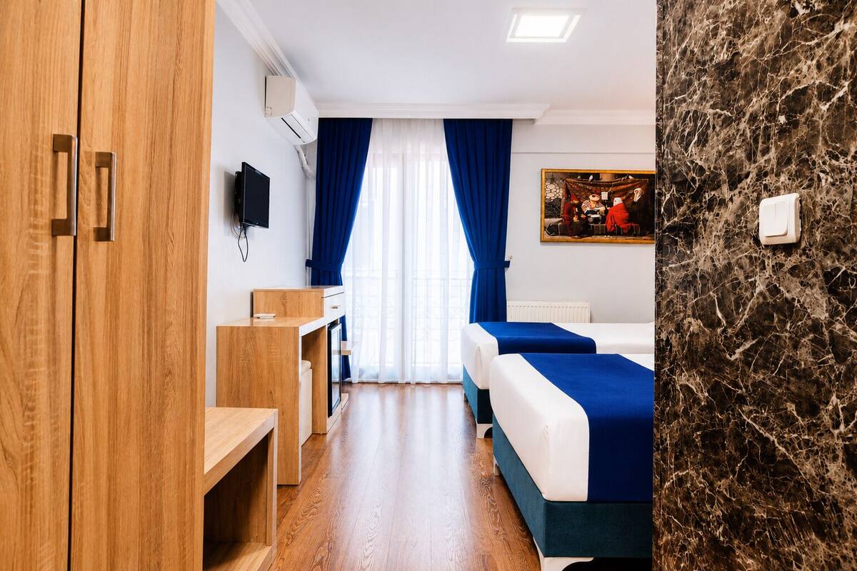 Quarto de hotel com guarda-roupa de madeira, escrivaninha, cama com roupa de cama azul, pintura na parede e parede com textura de mármore.
