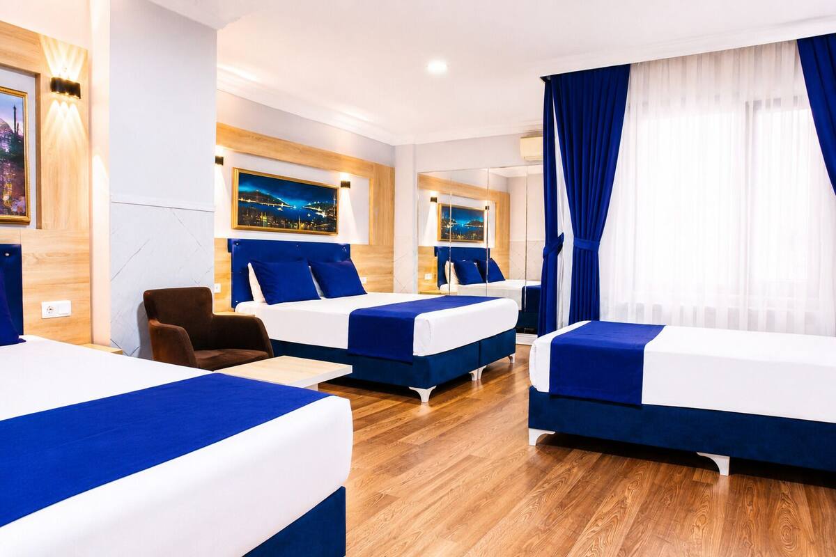 Quarto de hotel com duas camas, uma cadeira, uma escrivaninha e uma janela grande com cortinas.
