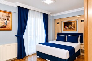 Een slaapkamer met een groot bed, een raam met blauwe gordijnen, een radiator en twee schilderijen aan de muur.