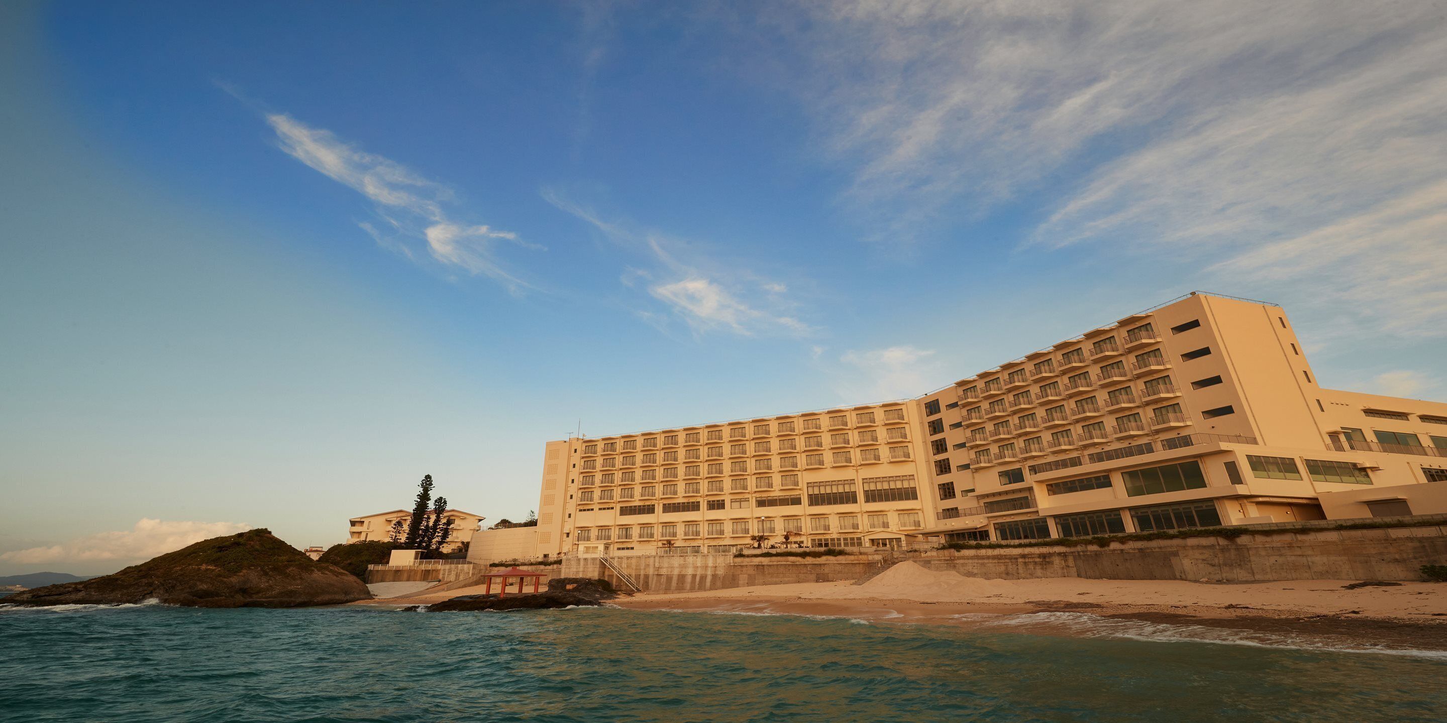 A large hotel with multiple floors, a beach in the foreground, and a rocky outcrop in the background.
