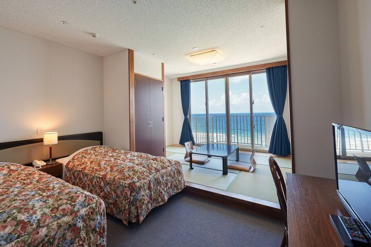A hotel room with two beds, a desk with a computer, a chair, and a balcony with a view of the ocean.