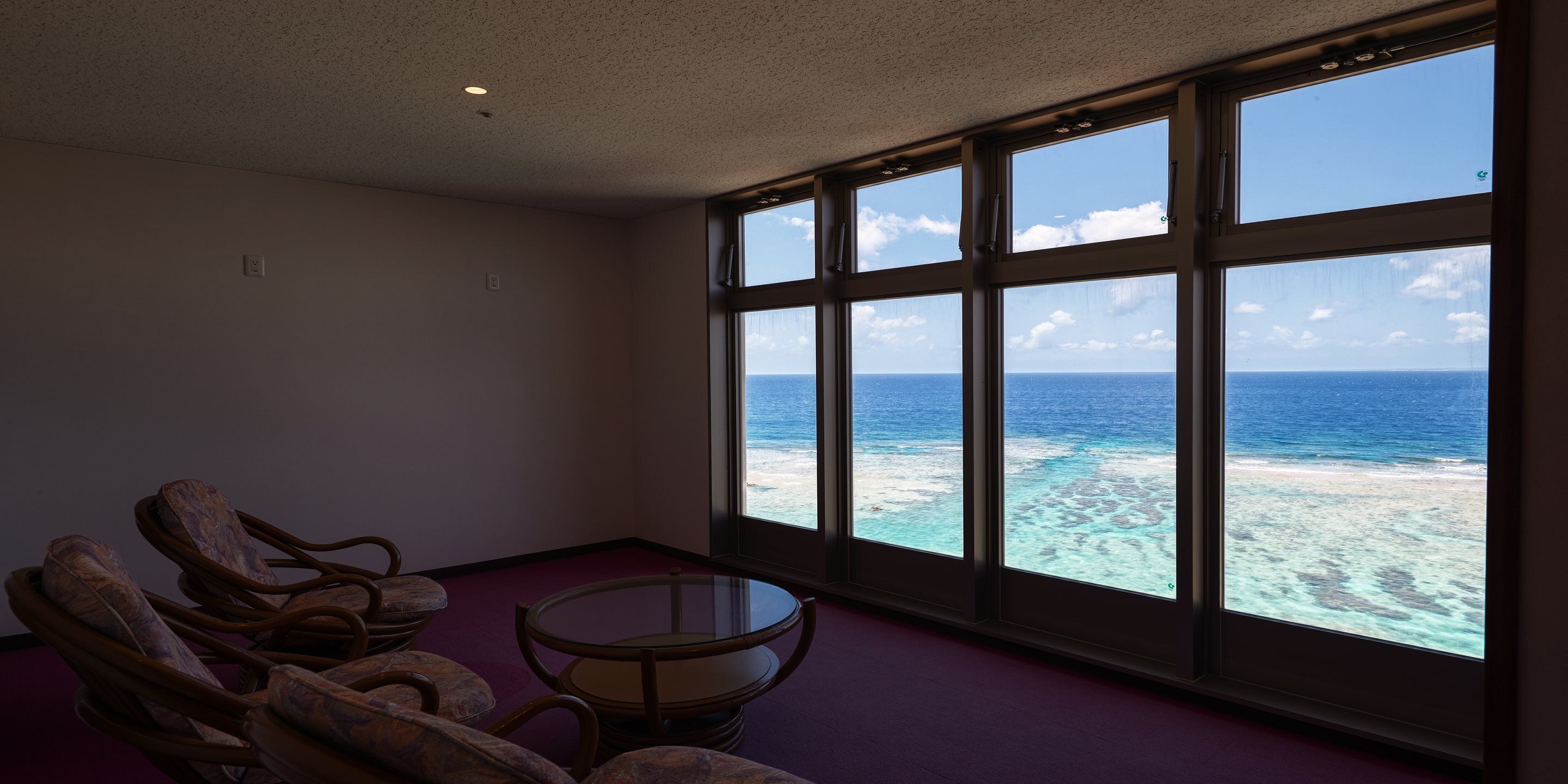 Ein Zimmer mit großem Fenster und Blick auf Ozean und Strand. Korbstühle und ein kleiner Tisch sind vorhanden.