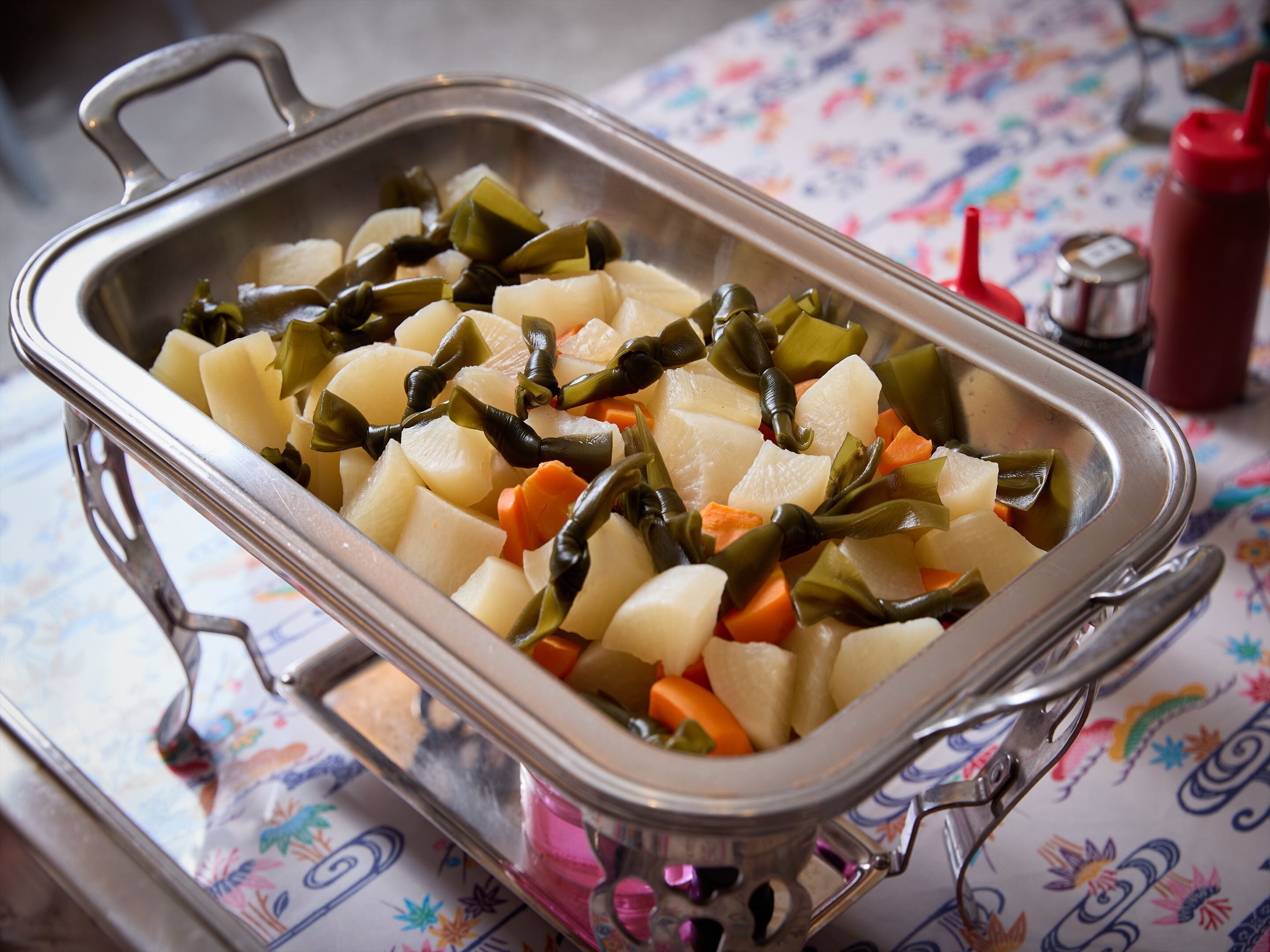 A stainless steel serving dish with cubed potatoes, carrots, and green vegetables on a table with a floral tablecloth.