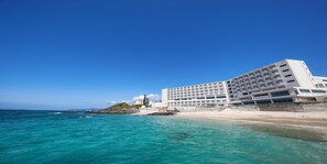 A coastal hotel with a modern design, featuring multiple floors and balconies, situated next to a sandy beach and the ocean. - Hotel Miyuki Beach (Onna)