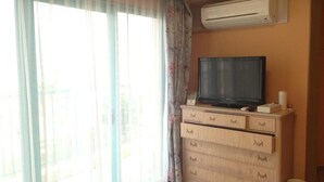 A room with a television on a wooden cabinet, a window with sheer curtains, and an air conditioning unit on the wall. - Hotel Bell Harmony Ishigakiisland (Ishigaki)
