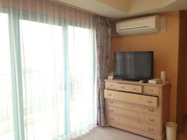 A room with a television on a wooden cabinet, a window with sheer curtains, and an air conditioning unit on the wall.