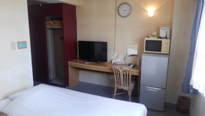A hotel room with a bed, a desk with a computer, a chair, a microwave, and a refrigerator.