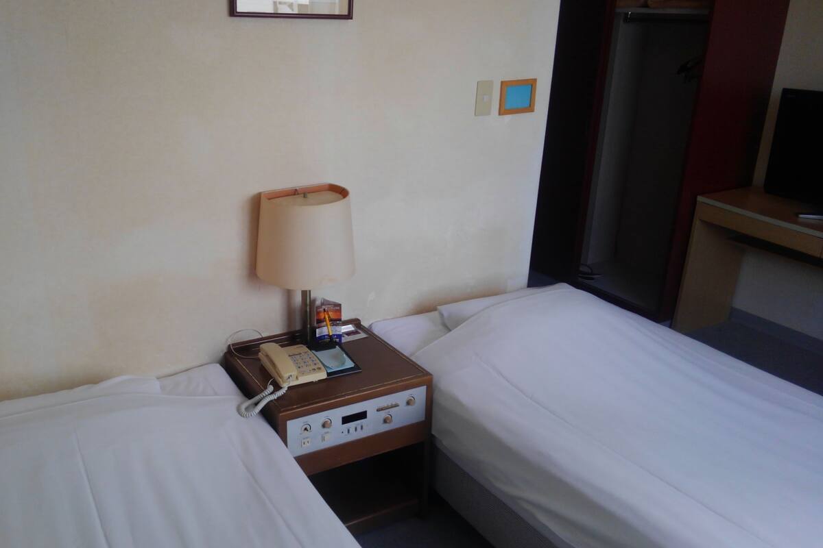 Habitación de hotel con dos camas, una mesita de noche con lámpara, teléfono y una pequeña estantería.