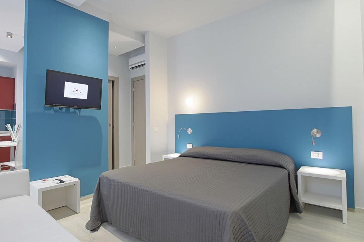 Ein modernes Hotelzimmer mit einem großen Bett, einem an der Wand befestigten Fernseher und einem kleinen Nachttisch mit Lampe.