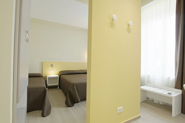 A hotel room with two single beds, a bedside table with a lamp, a bench by the window, and a wall-mounted light fixture.