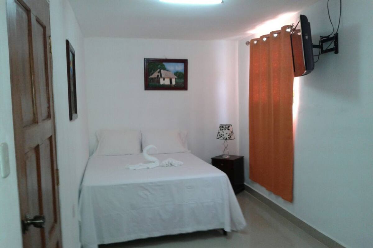Ein Schlafzimmer mit einem Bett, einem Nachttisch, einer Lampe, einem Gemälde und einem an der Wand befestigten Fernseher.