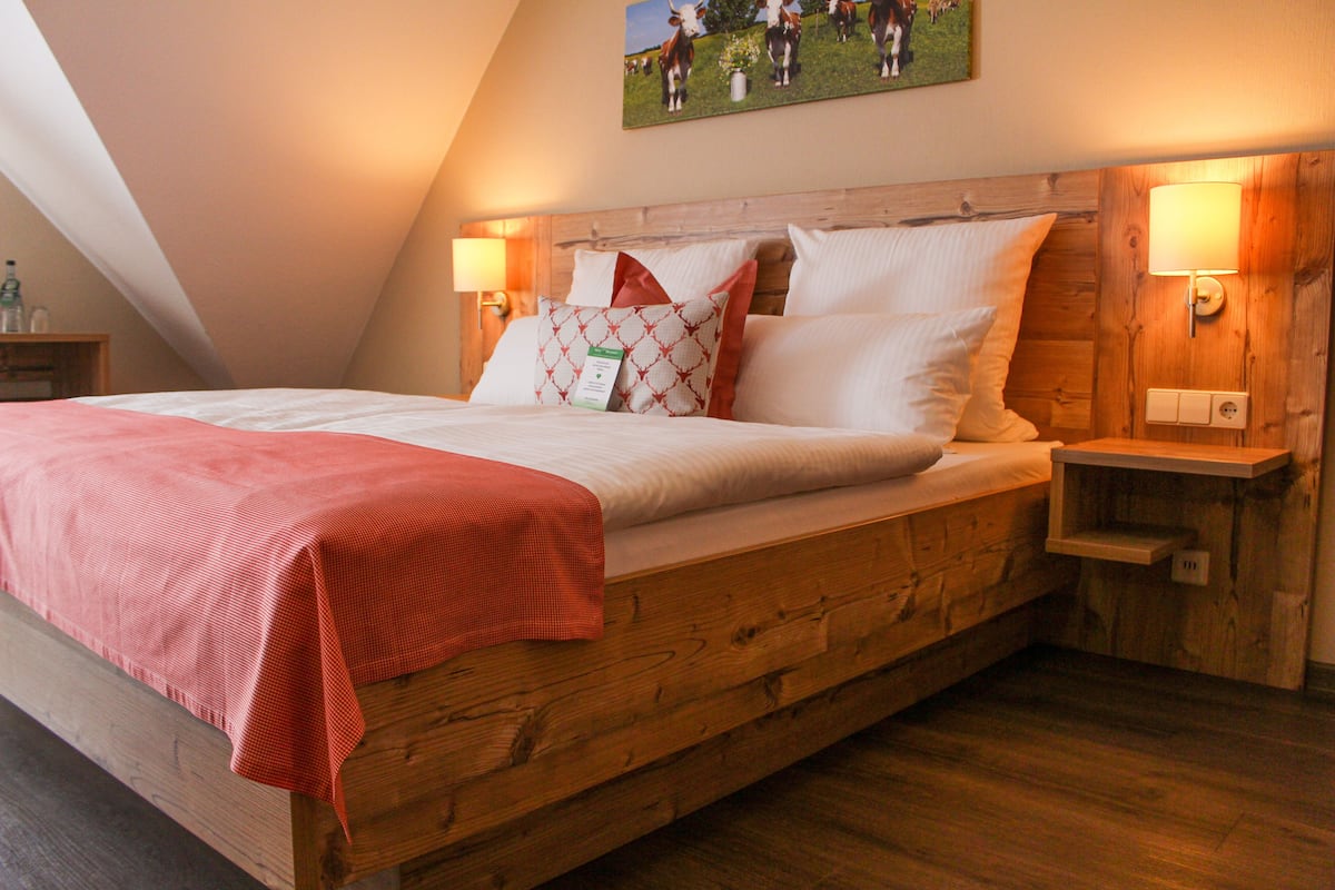 A bedroom with a wooden bed, a red blanket, two bedside lamps, and a painting on the wall.