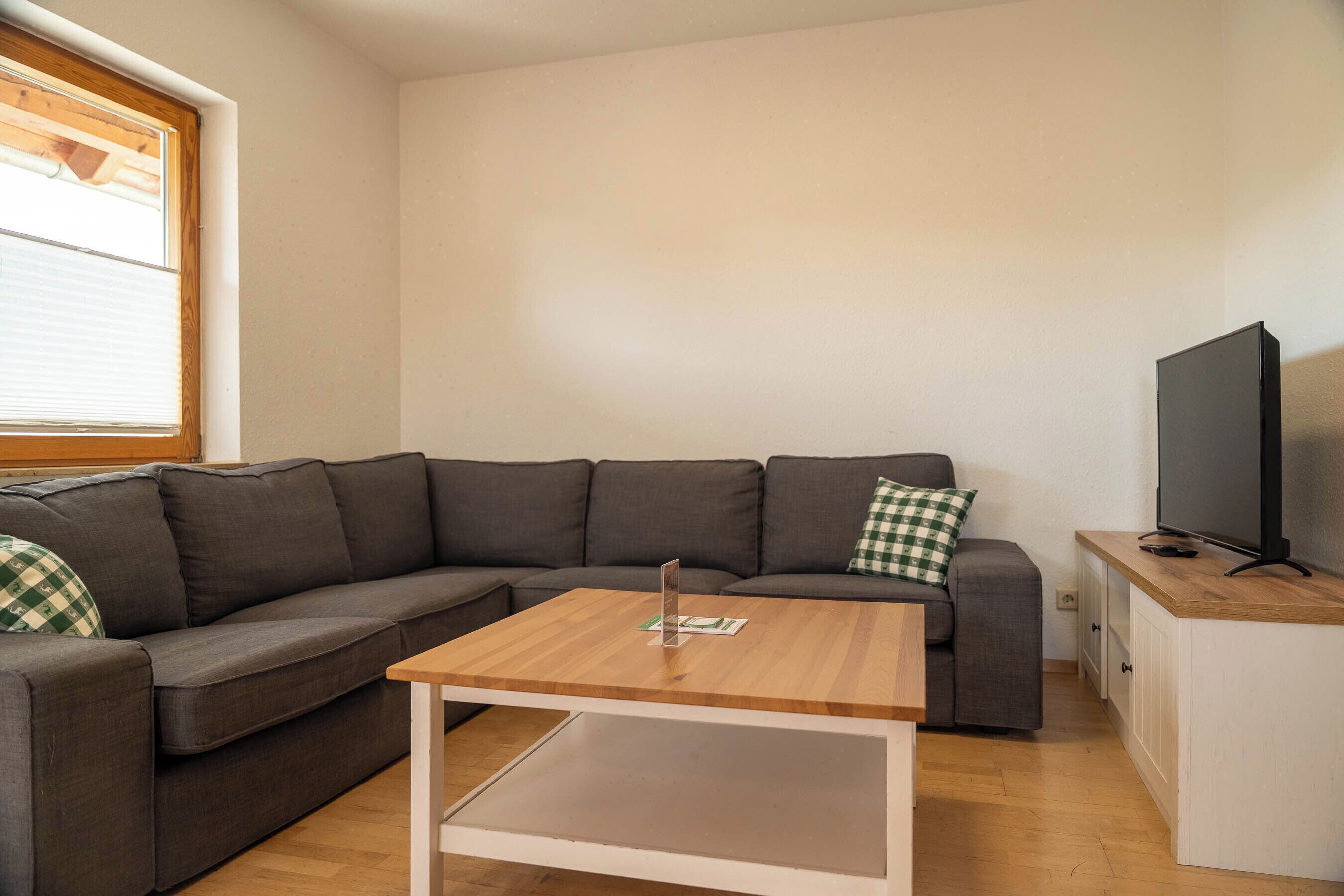 Una sala de estar moderna con un sofá seccional, una mesa de centro de madera, un televisor sobre un mueble bajo y una ventana con persianas.