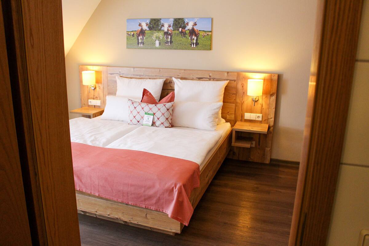 A neatly made bed with a wooden headboard, two bedside tables with lamps, and a painting of cows on the wall.
