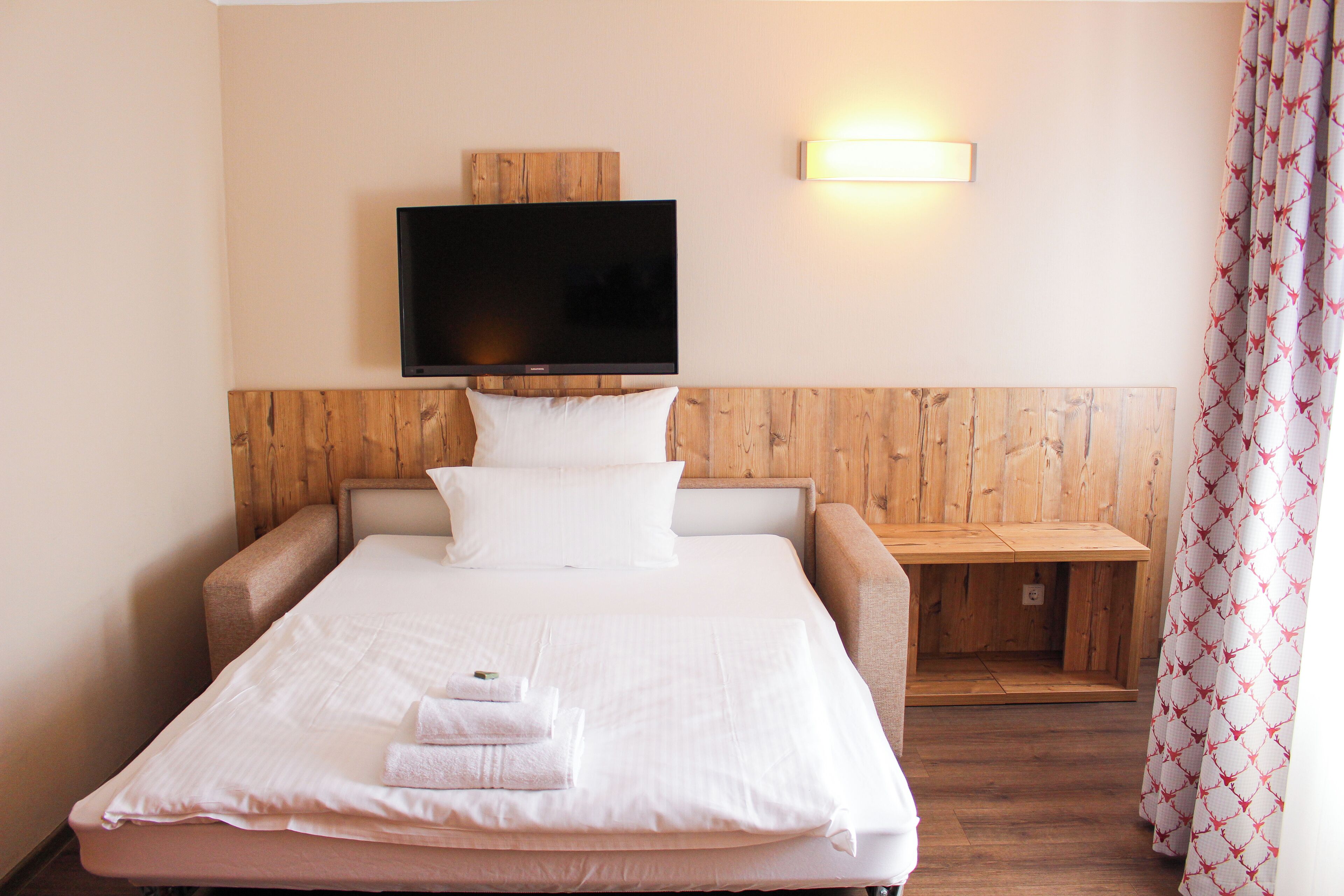 Cama bem arrumada com lençóis brancos e uma toalha dobrada, cabeceira de madeira, televisão montada e mesa de cabeceira com abajur.
