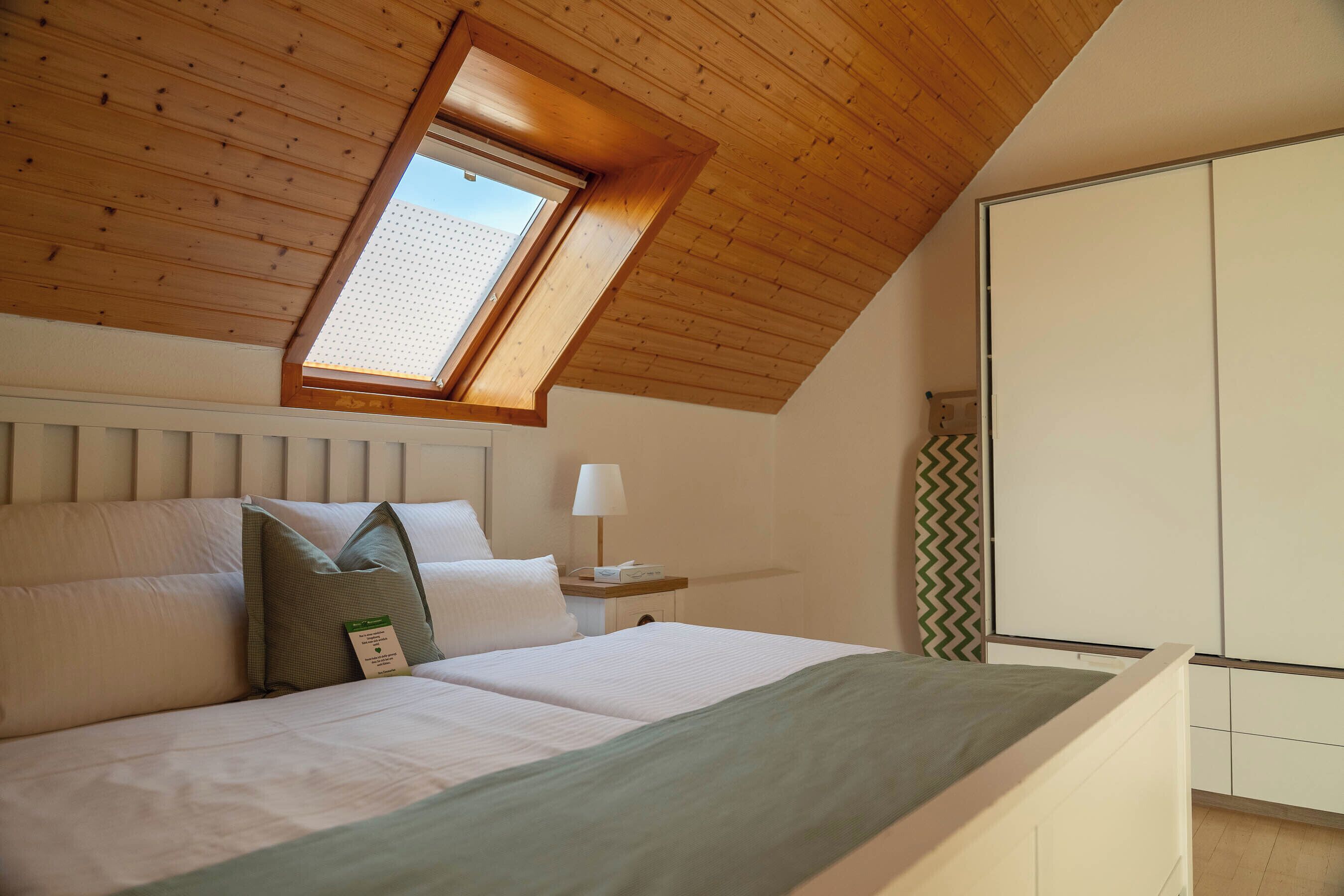 Ein Schlafzimmer mit schrägverlegtem Holzboden, einem Oberlicht, einem Bett mit weißer Bettwäsche, einem Nachttisch mit Lampe und einem Kleiderschrank.