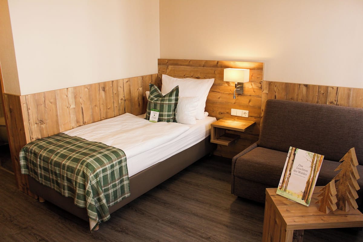 A neatly made bed with a plaid blanket, a wooden headboard, a nightstand with a lamp, and a brown armchair.