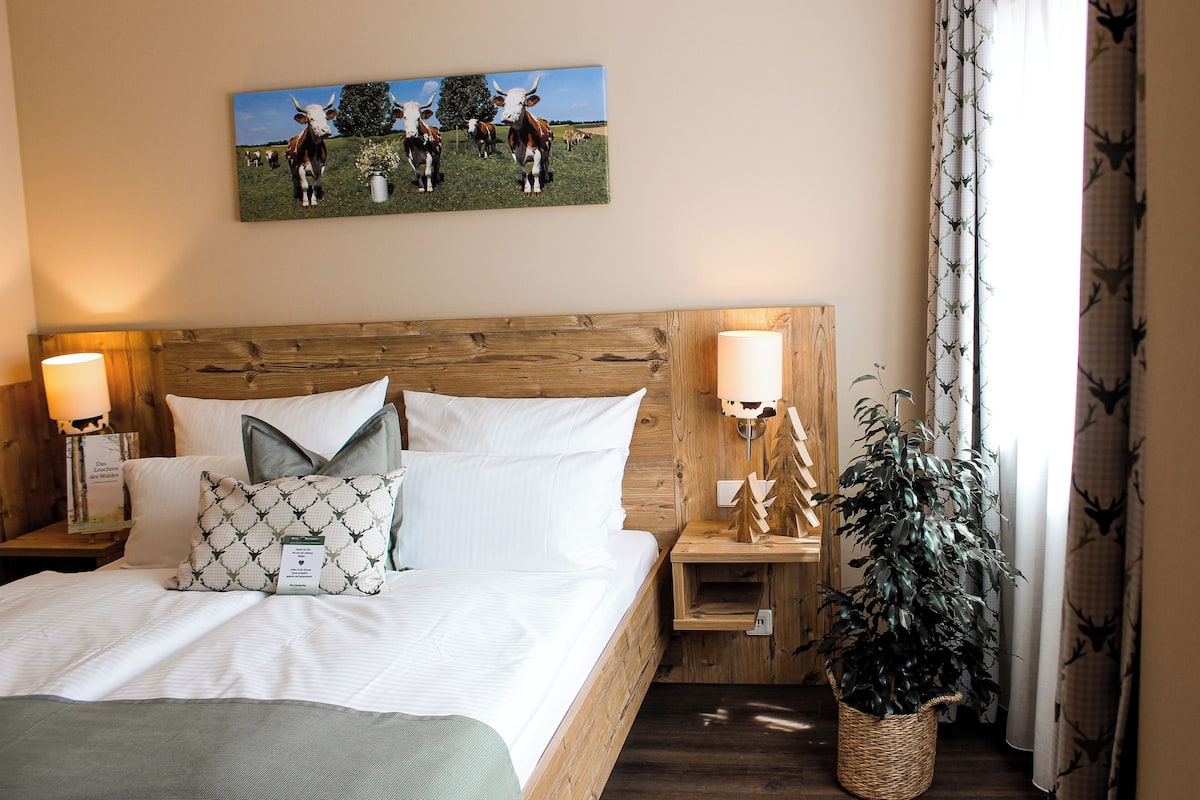 A hotel room with a wooden headboard, a painting of cows, a bedside lamp, a bedside table, a potted plant, and a window with curtains.