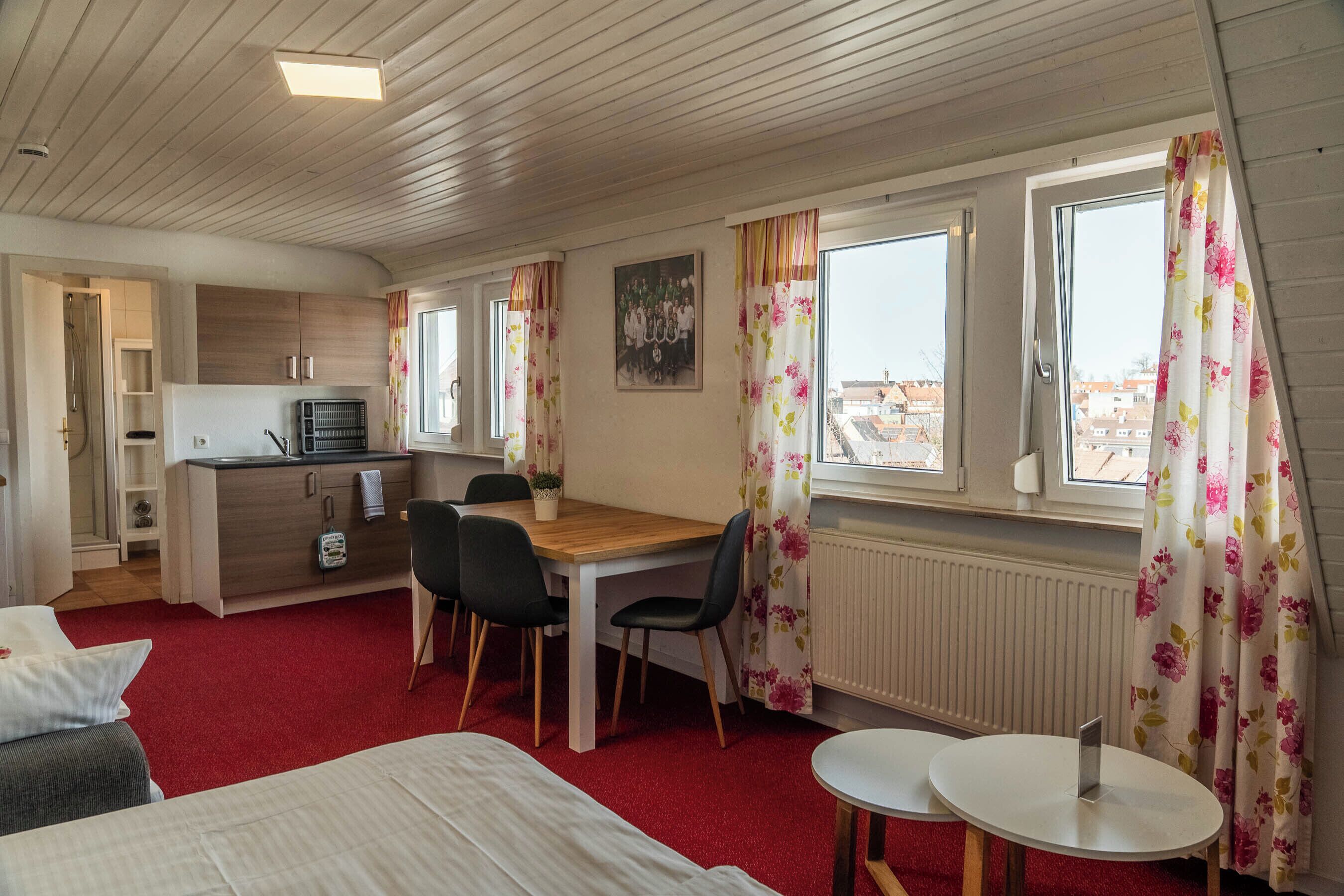 Una habitación con una cama, una mesa de comedor con sillas, una pequeña cocina y una ventana con cortinas.