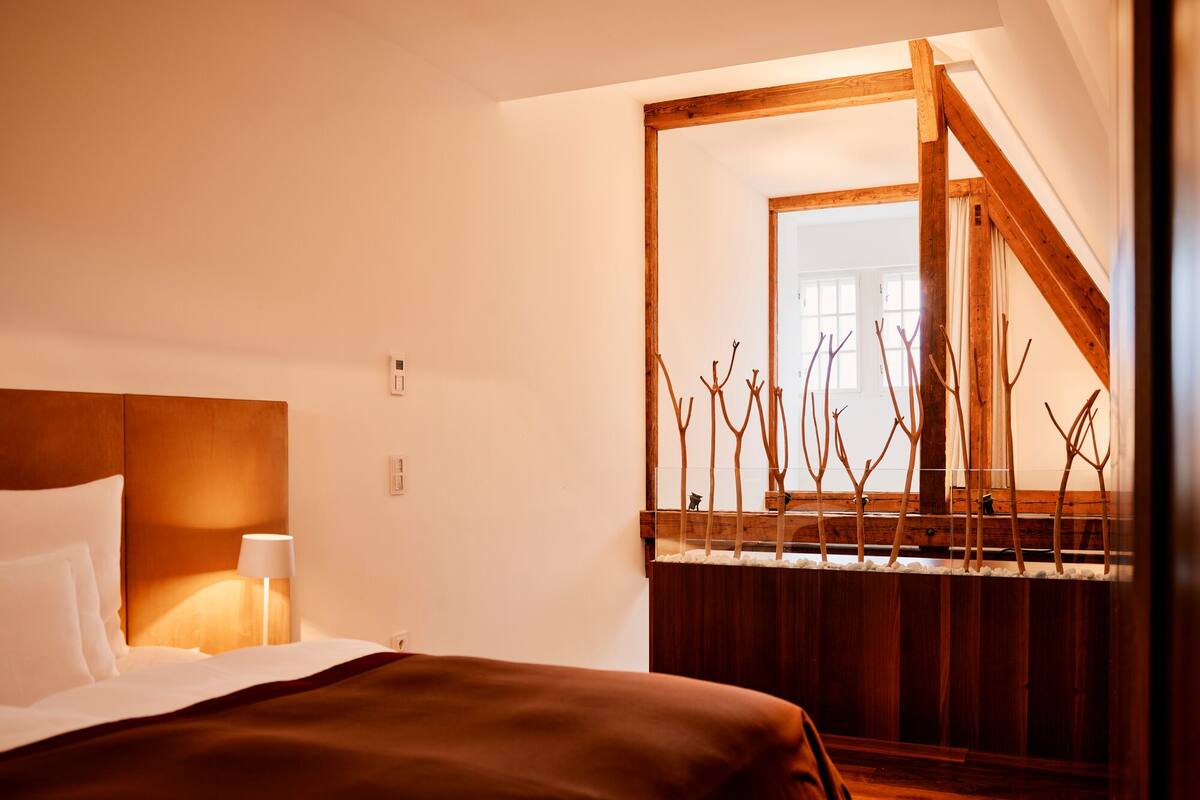 A hotel room with a bed, a nightstand, a lamp, and a wooden staircase leading to a balcony.