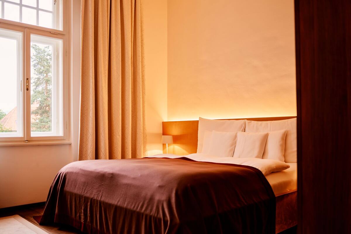 A hotel room with a bed, a window, curtains, and a nightstand with a lamp.