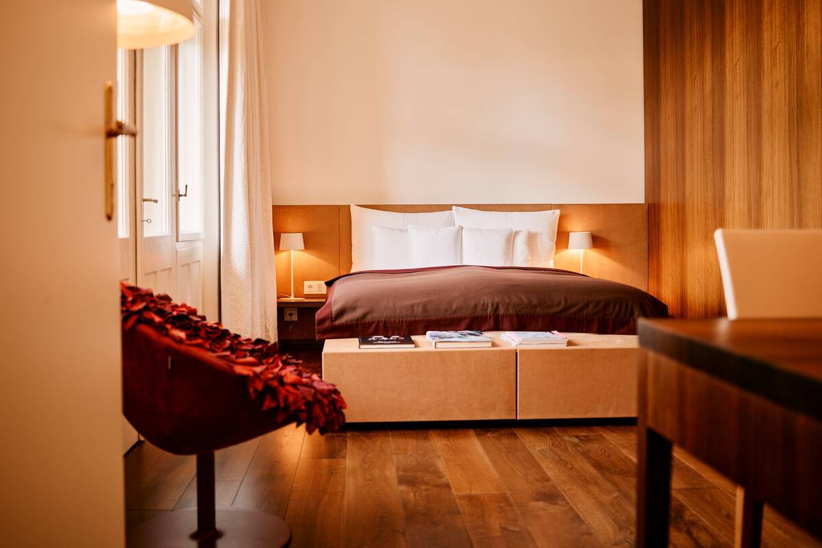 A hotel room with a bed, bedside lamps, a wooden floor, and a red chair.