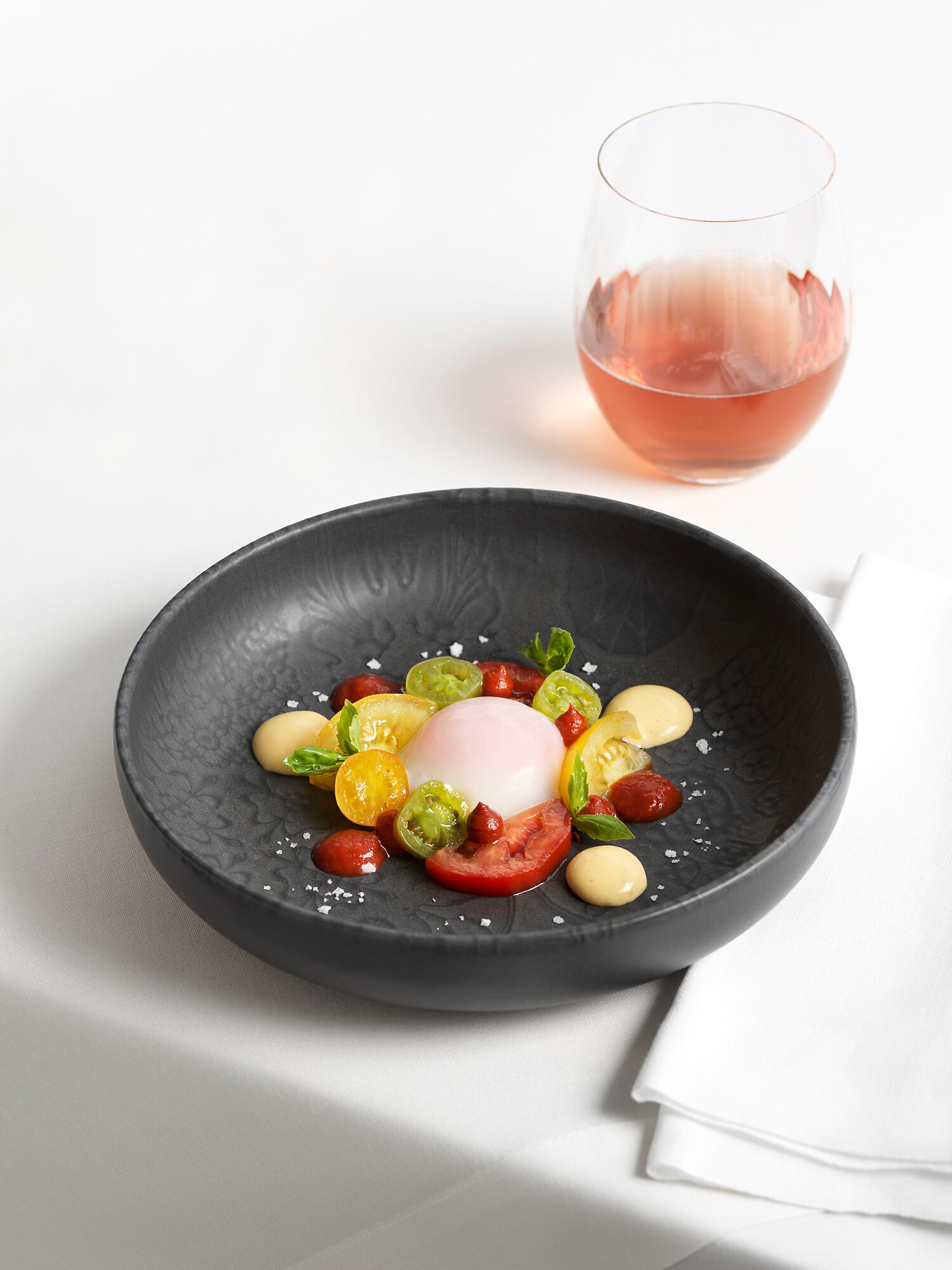 Ein schwarzer Teller mit pochiertem Ei, Cocktailtomaten, Oliven und Basilikum auf einem weißen Tisch.