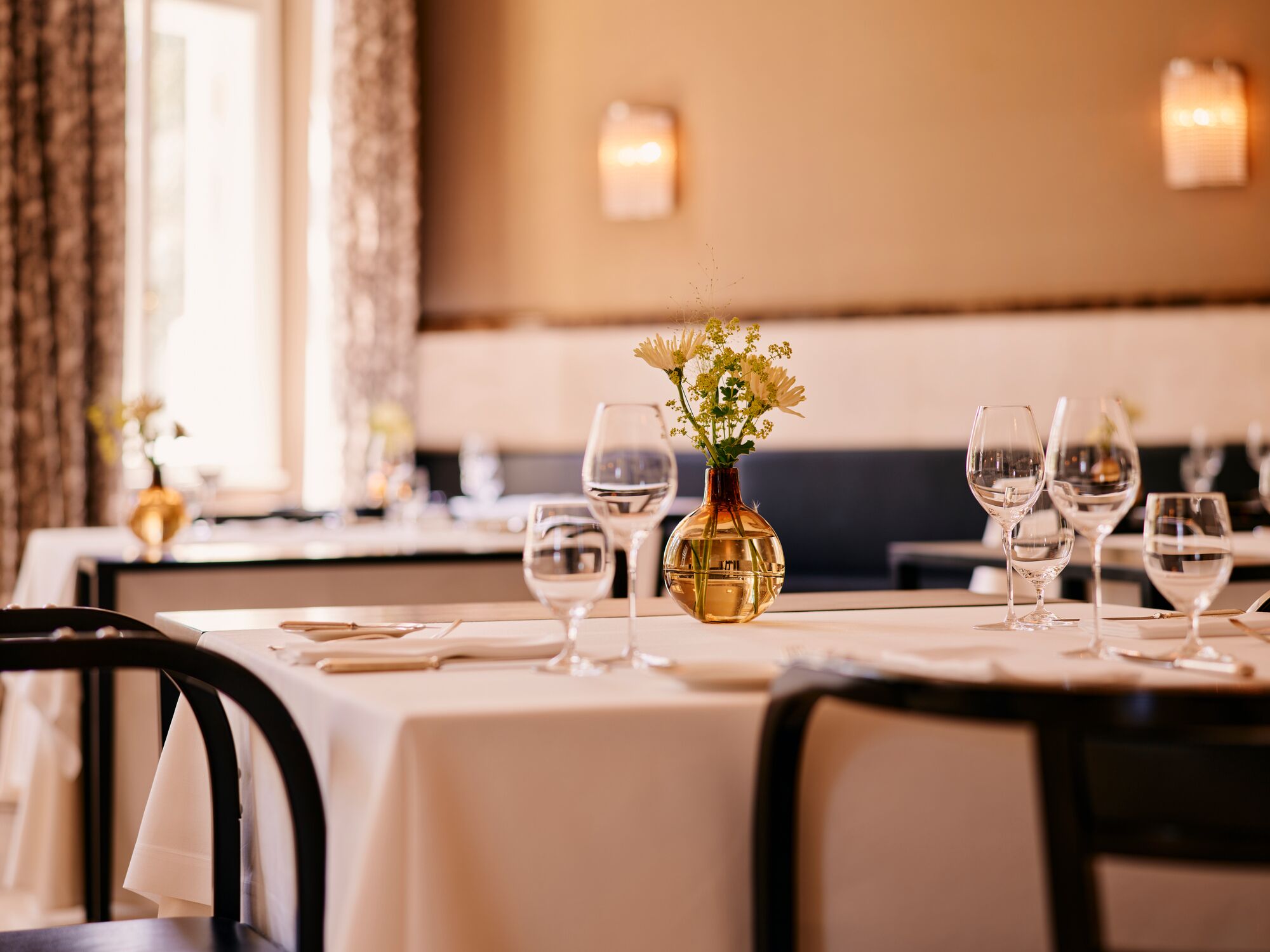 A dining table set with white tablecloths, wine glasses, and a vase with flowers.
