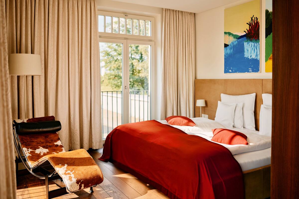 A hotel room with a bed, a chair, a painting, and a window with curtains.