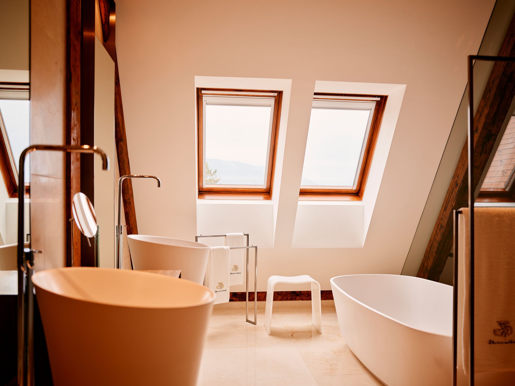 Une salle de bain moderne avec une baignoire sur pieds, une baignoire plus petite et un espace douche. Deux fenêtres laissent entrer la lumière naturelle et offrent une vue sur l’extérieur.