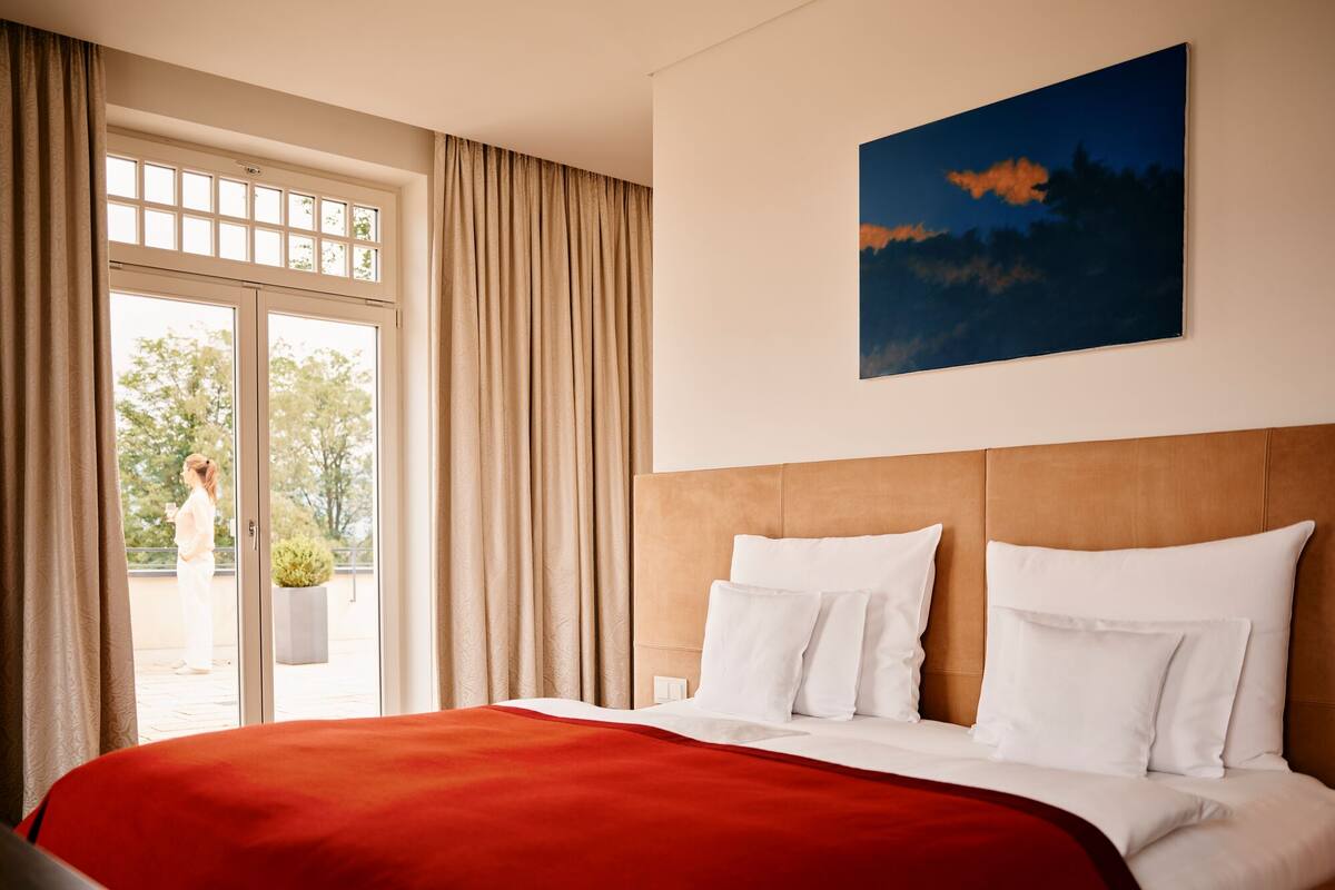A hotel room with a large bed, a red blanket, white pillows, a wooden headboard, and a painting of a sunset on the wall.