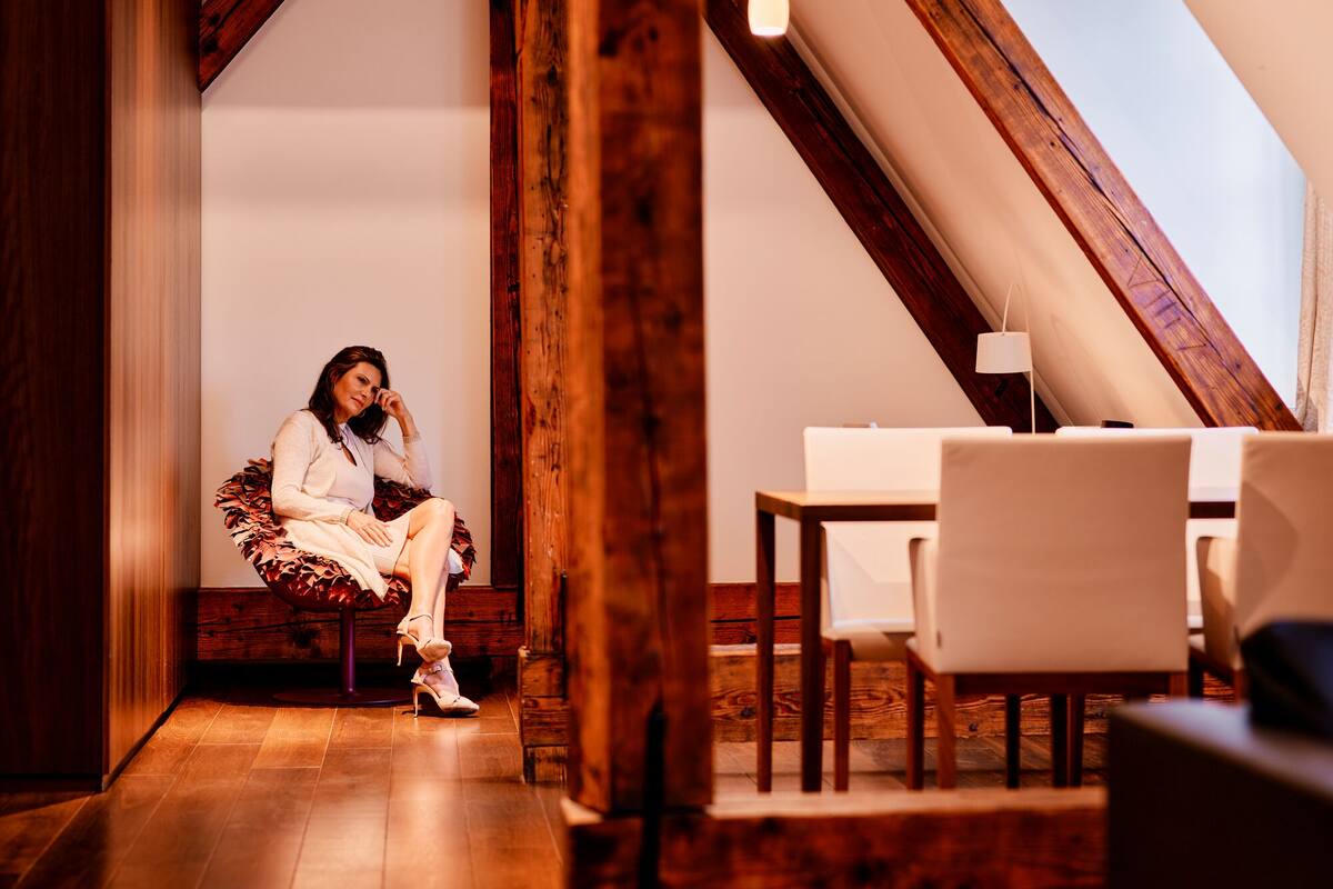 A woman sitting on a chair in a room with wooden beams, a table, and a hanging light fixture.