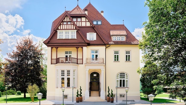 Un edificio de dos pisos con estilo arquitectónico tradicional, que destaca por una entrada imponente, un balcón y un jardín ajardinado con senderos y plantas en macetas.