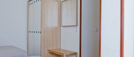 A bedroom with a bed, a wooden wardrobe, a small wooden table, and a mirror on the wall.