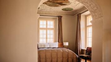 A bedroom with a large bed, two windows with curtains, a wooden nightstand, and a decorative ceiling.