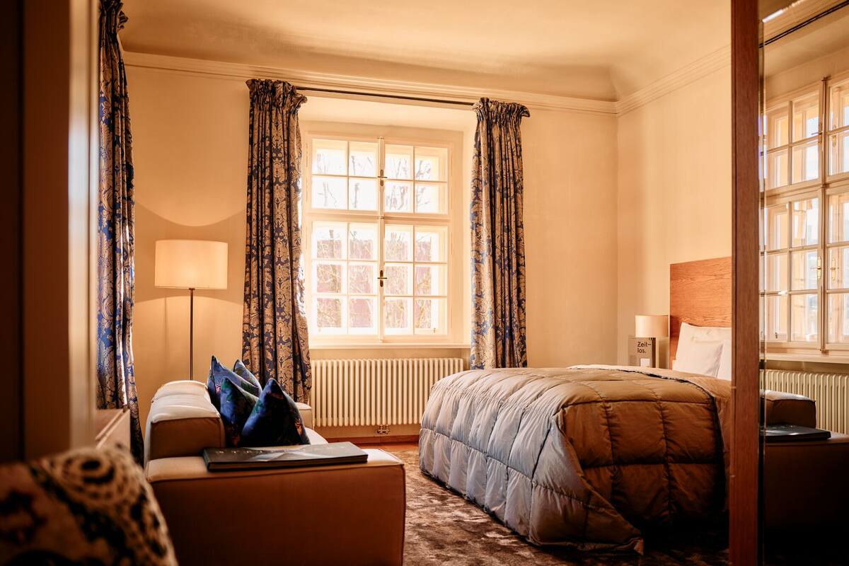 A hotel room with a bed, a desk, a chair, a lamp, and a window with curtains.