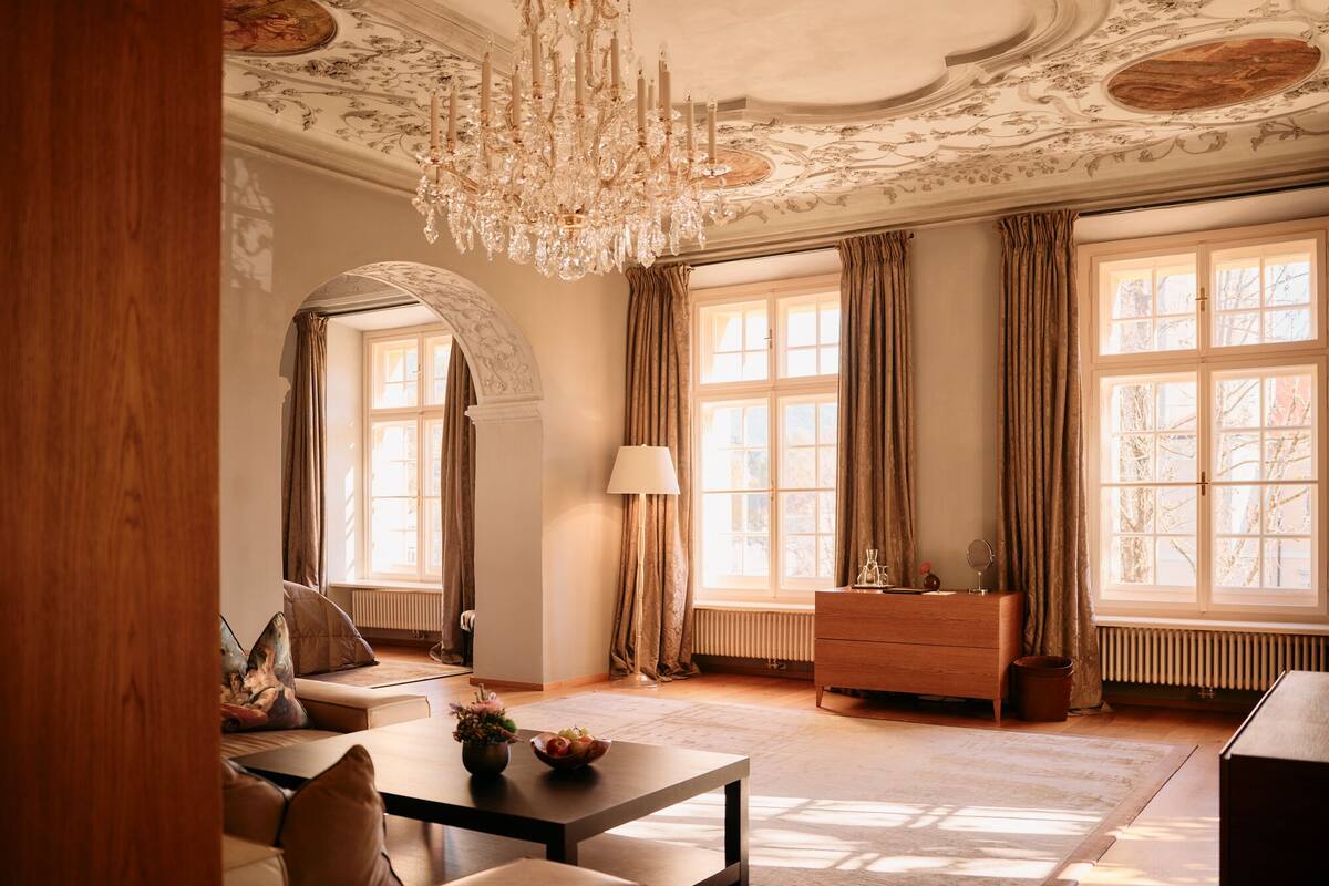 A spacious living room with a chandelier, large windows, a sofa, a coffee table, and a wooden cabinet.
