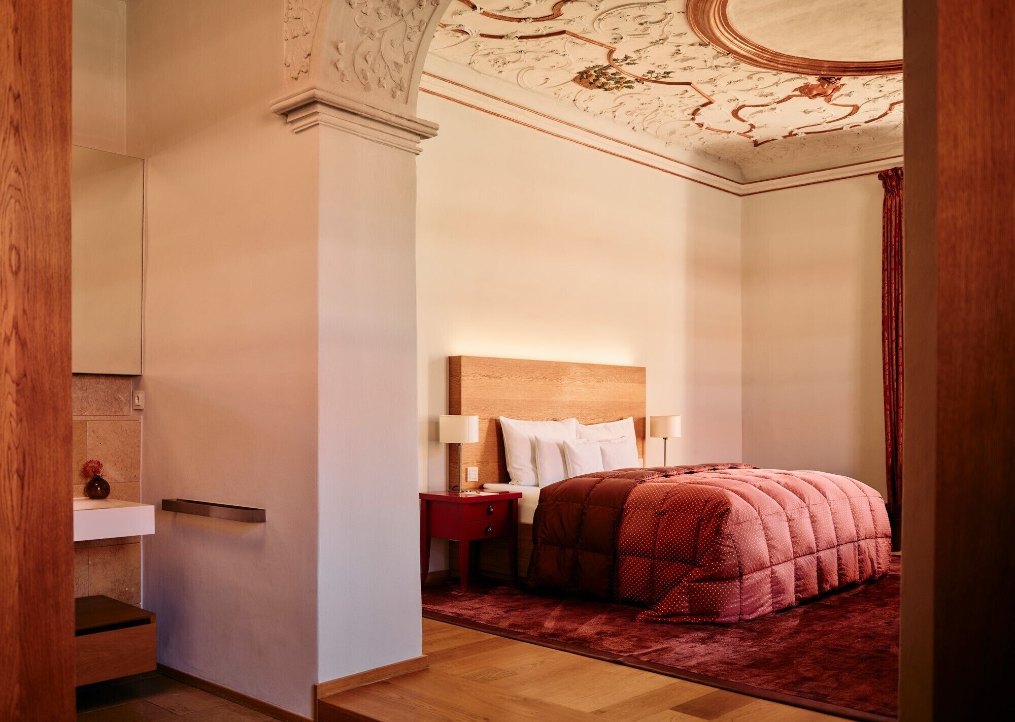 A hotel room with a wooden headboard, a red bedspread, and a nightstand with lamps.
