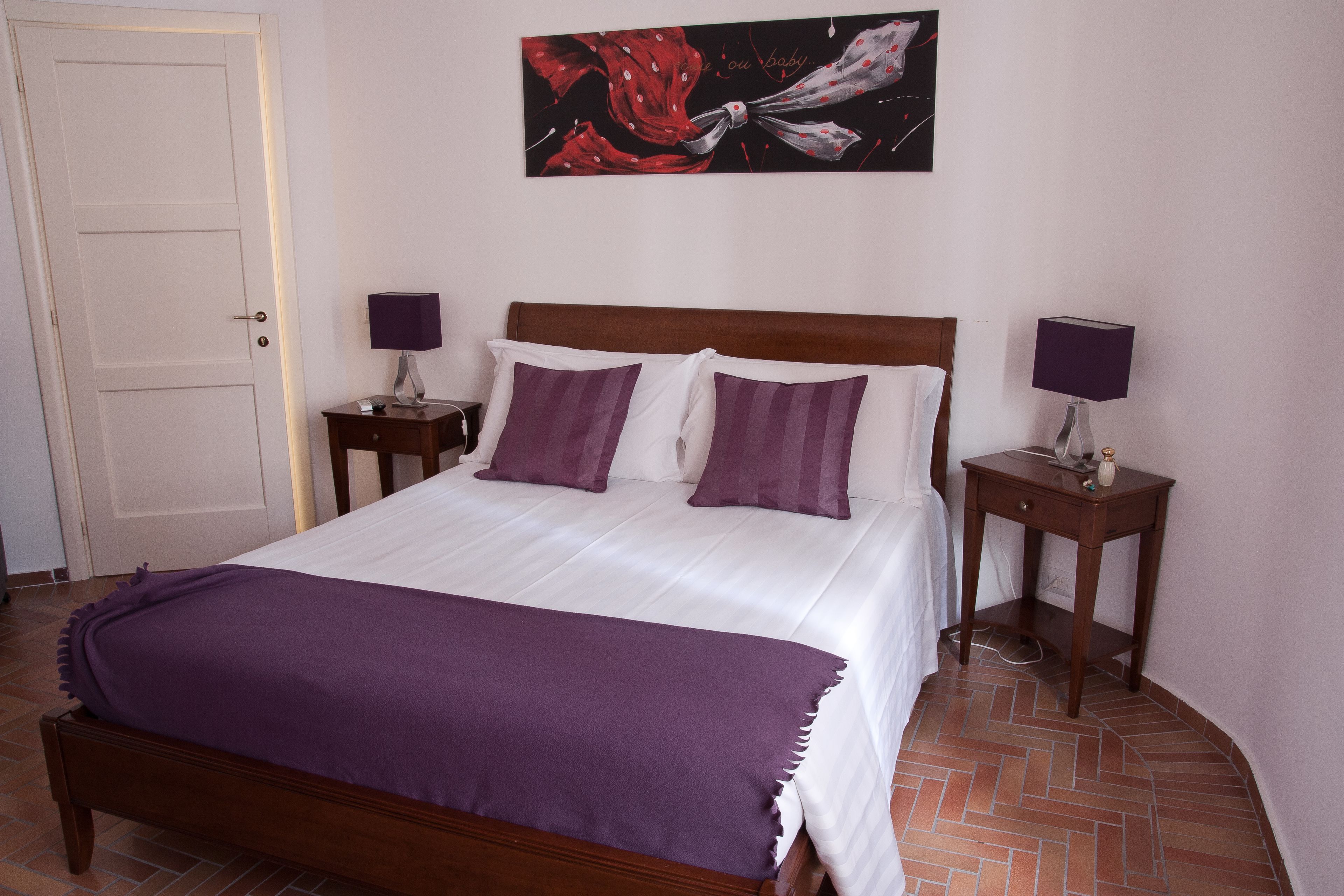 A neatly made bed with a purple blanket, two bedside tables with lamps, a wooden headboard, and a painting on the wall.